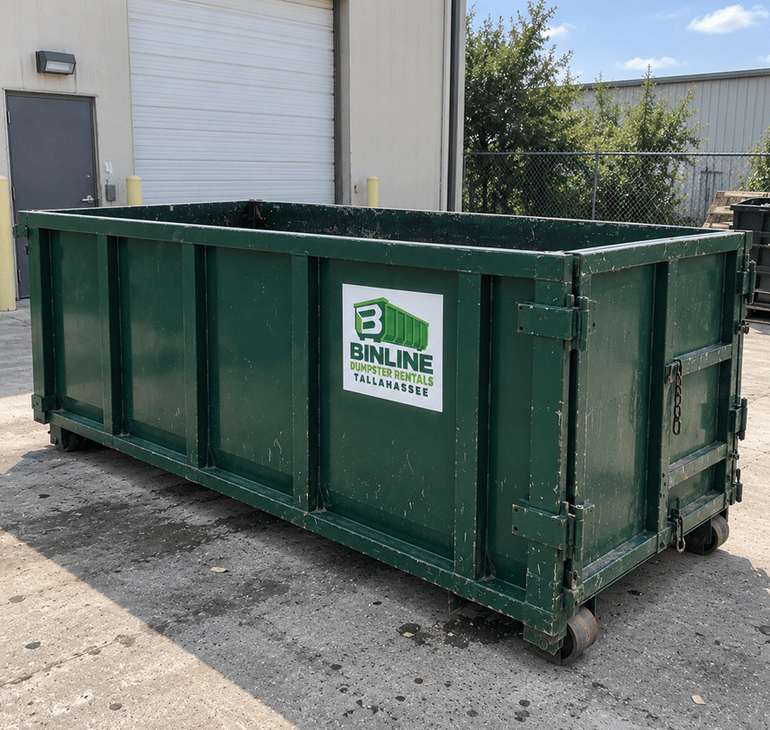 Green open-top roll-off dumpster with the Binline Dumpster Rentals Tallahassee logo on the side, parked on concrete beside a fence